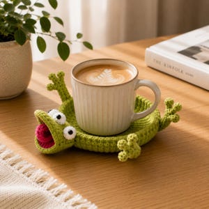 May include: A light green crocheted coaster shaped like a frog with a wide open mouth, white and black eyes, and four small feet, holding a beige ceramic mug filled with coffee featuring a white leaf-shaped foam design on the surface.