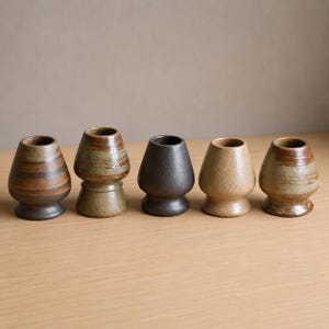 May include: A set of five ceramic matcha whisk holders arranged in a row. Each holder features a distinct finish, including brown horizontal stripes, mottled green and brown, solid matte black, light tan, and a variegated brown and green glaze.