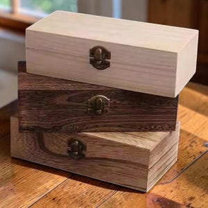 Peut inclure: Une pile de trois boîtes rectangulaires en bois avec des couvercles à charnières et des fermoirs en métal couleur bronze. La boîte supérieure est beige clair, celle du milieu est brun foncé avec un grain visible, et la base est brun moyen.