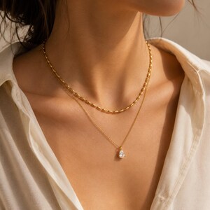 Könnte beinhalten: Ein geschichtetes Goldhalsketten-Set bestehend aus zwei verschiedenen Ketten. Die obere Kette zeigt eine Reihe kleiner, länglicher Goldperlen, während die untere Kette eine feine Gliederkette mit einem einzelnen birnenförmigen, klaren Edelsteinanhänger ist.