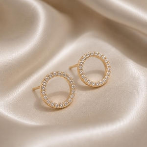 May include: A pair of gold-toned stud earrings featuring a circular design. Each earring is encrusted with a series of small, clear, sparkling stones set along the entire circumference of the ring, creating a bright and reflective appearance.
