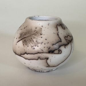 May include: A small, round ceramic vase with a matte white finish. The surface features dark, smoky black markings, including a distinct feather imprint on the upper left side and fine, irregular speckles scattered across the rounded body.