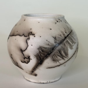 May include: A round ceramic vase with a matte white finish, featuring dark charcoal-colored smoke-fired markings. The surface displays a prominent black feather-like impression along with fine, irregular crackle lines and scattered dark speckles.