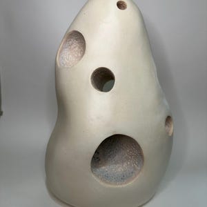 May include: An off-white ceramic sculpture with an irregular, rounded shape featuring five circular openings of varying sizes. The interior surfaces of the holes show a textured, speckled finish, while the exterior surface is smooth and matte.