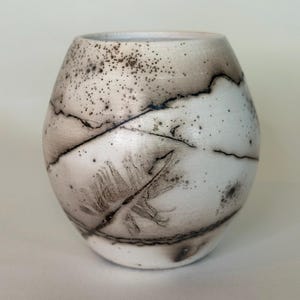May include: A round ceramic vase with a white base and a matte finish. The surface features dark brown and black smoke-fired markings, including thin, jagged lines and a prominent feather-like imprint on the front, with scattered dark speckles.