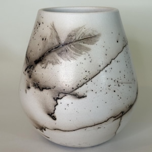 May include: A white ceramic vase with a rounded, tapered shape featuring a black feather imprint on the upper side. The surface is decorated with fine black crackle lines and dark speckles that create a textured, organic appearance throughout.