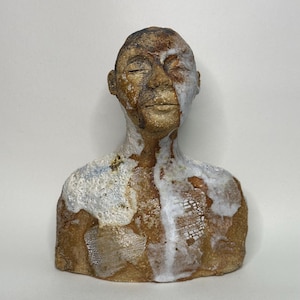 Può includere: Un busto in ceramica di una testa e spalle umane con una finitura marrone terrosa e ruvida. La scultura presenta macchie di smalto bianco che sembrano colare sul viso e sul petto, rivelando intricate impronte a griglia sulla superficie.