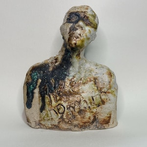 Könnte beinhalten: Eine Keramikbüste mit einer strukturierten, cremeweißen Oberfläche und gesprenkelten braunen, grünen und schwarzen Glasurakzenten. Die Figur wirkt verwittert, mit sichtbaren Rissen und eingravierten Buchstaben im Brustbereich, was ihr eine rohe Optik verleiht.