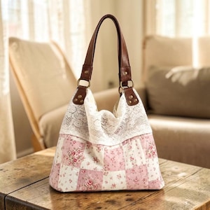 May include: A hobo-style handbag featuring a patchwork design of pink and white floral fabric squares. The upper section is adorned with a wide band of white lace. The bag includes two brown leather straps attached with gold-tone metal rings.
