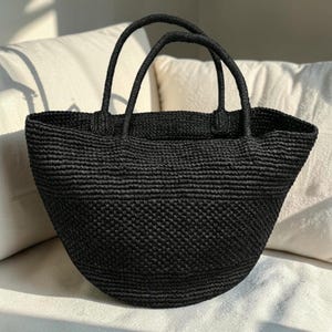 Può includere: Una borsa tote nera caratterizzata da una struttura intrecciata e testurizzata, con un'ampia apertura superiore e due manici arrotondati e robusti. La borsa presenta una base curva a semicerchio e un motivo a punti denso e uniforme.