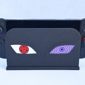 Pode incluir: Uma base de carregamento para consola de videojogos preta com um gráfico de dois olhos distintos, um com um padrão Sharingan vermelho e o outro com um padrão Rinnegan roxo. Os comandos pretos têm capas de joystick com pegadas de pata.