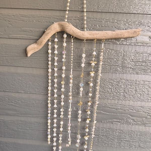 May include: A wall hanging decoration featuring a curved piece of light brown driftwood suspended by a pearl chain. Seven vertical strands of clear, iridescent, and white beads, along with small gold-toned metal angel charms, hang from the wood.