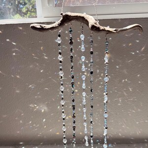 May include: A hanging mobile featuring a piece of natural driftwood at the top with five dangling strands of beads. The strands consist of clear, faceted glass crystals, metallic silver-toned spacers, and small blue beads, creating light reflections.