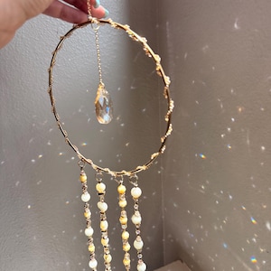 May include: A gold-toned metal hoop suncatcher featuring a central clear teardrop crystal pendant suspended from a delicate chain. Four vertical strands hang from the bottom of the hoop, decorated with alternating cream-colored beads and small clear crystals.