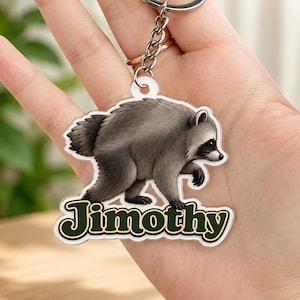 May include: A clear acrylic keychain featuring a detailed illustration of a gray and black raccoon walking. Below the raccoon, the name Jimothy is printed in a bold, dark green font with a white outline. The charm is attached to a silver metal ring.