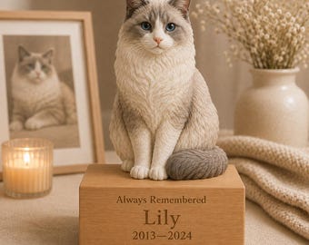May include: A cat figurine with cream and grey fur and blue eyes sits on a rectangular wooden base. The base is engraved with the text Always Remembered, the name Lily, and the years 2013-2024. The cat has a textured coat and a curled tail.