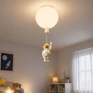 May include: A ceiling light fixture featuring a large, spherical, frosted white glass shade attached to a white and gold astronaut figurine. The astronaut is suspended by a thin metal rod, appearing to hold the glowing orb above its helmet.