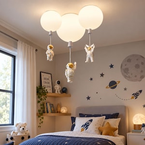 May include: A ceiling light fixture featuring three white spherical glass globes suspended by rods. Each rod holds a small white and gold astronaut figurine. The astronauts are positioned as if floating, with one waving and two hanging vertically.