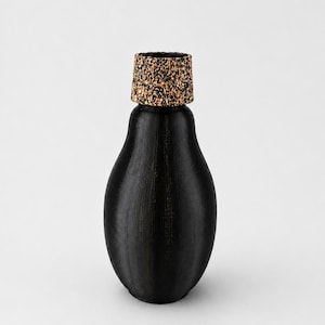 May include: A pear-shaped vase with a matte black finish and a textured, vertical ribbed surface. The top opening is fitted with a cylindrical stopper featuring a speckled, natural brown cork-like material with dark flecks throughout the surface.