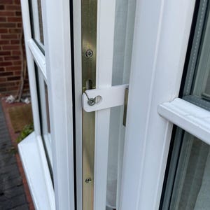 May include: A white plastic window restrictor latch mounted on a white uPVC window frame. The mechanism features a flat, rectangular arm secured with a metal screw, designed to limit the opening distance of the window sash against the frame.