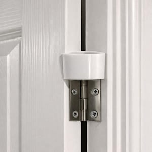 May include: A white plastic door hinge pin bumper mounted on a silver metal door hinge. The hinge is attached to a white panelled door and frame with four visible screws. The bumper features a cylindrical top section and a flat base plate.