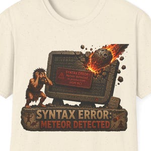 May include: A cream-colored t-shirt featuring a graphic of a caveman reacting to a meteor striking a stone computer monitor. The text reads SYNTAX ERROR: METEOR DETECTED, with a smaller red box on the screen displaying SYNTAX ERROR - Meteor detected.