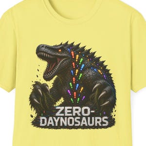 May include: A pale yellow short-sleeve t-shirt featuring a graphic of a dark, textured dinosaur with an open mouth. A trail of colorful exclamation points flows from the dinosaur's neck. The text "ZERO-DAYNOSAURS" is printed in white below.