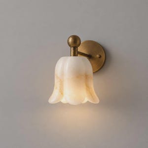 May include: A wall sconce featuring a bell-shaped, translucent white and beige marble shade with a smooth, scalloped edge. The fixture is supported by a circular brass mounting plate and a short, cylindrical arm with a spherical top detail.