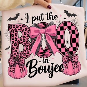 Peut inclure: Un t-shirt rose pâle avec le texte noir I put the BOO in Boujee. Le motif comprend une lettre B à imprimé léopard rose, un O camouflage rose, un O à carreaux roses, deux citrouilles roses avec des nœuds et des chauves-souris.