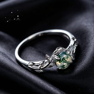 May include: A silver-toned ring featuring a round, faceted moss agate gemstone with green inclusions. The band is designed with a detailed relief of leaves and vines extending from the setting, which holds the stone in a four-prong mount.