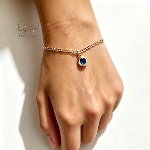 May include: A gold chain bracelet featuring a single circular charm with a deep blue stone set in the center. The bracelet has a delicate link design and a polished metallic finish. The logo Gem Atelier is visible in the upper left corner.