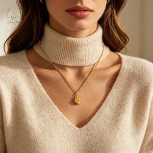 May include: A gold-toned necklace featuring a small, detailed crown pendant suspended from a delicate beaded chain. The pendant is centered on the chest, resting against a beige ribbed knit sweater with a high turtleneck collar.