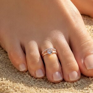 May include: A rose gold toe ring featuring a central heart-shaped stone flanked by smaller round stones. The band has a polished metallic finish and is worn on the second toe, showcasing a delicate design with clear, sparkling accents.