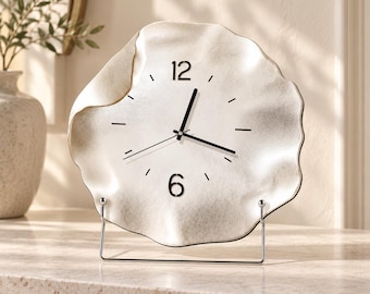 May include: A silver-toned table clock with an irregular, wavy edge and a textured metallic finish. The clock face features black hour and minute hands, a thin second hand, and black numerals at the 12 and 6 positions. It rests on a thin metal stand.