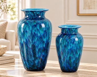 May include: Two glass vases featuring a vibrant, multi-tonal blue and teal feathered pattern. The larger vase stands to the left, while a smaller, matching vase is positioned to the right. Both vessels have a wide, flared rim and a smooth finish.