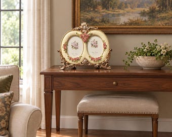 May include: A double oval picture frame with an ornate gold-toned border, featuring sculpted pink rose accents at the top and bottom of each frame. The frame has a cream-colored background and stands on decorative feet on a wooden table.