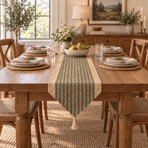 May include: A rectangular woven table runner featuring a central olive green striped pattern bordered by natural tan fibers, finished with a single tassel at the pointed end, displayed on a wooden dining table with place settings and a bowl of pears.