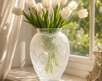 May include: A clear glass vase with an embossed floral pattern containing a bouquet of white tulips with green stems and leaves. The vase has a rounded body and a wide opening, reflecting light with an iridescent sheen while holding the flowers.