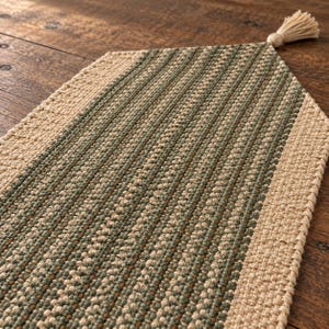 May include: A woven table runner featuring a central panel of sage green and beige textured fibers, bordered by a solid beige woven edge. The runner has a pointed end finished with a single beige tassel attached to the center point.