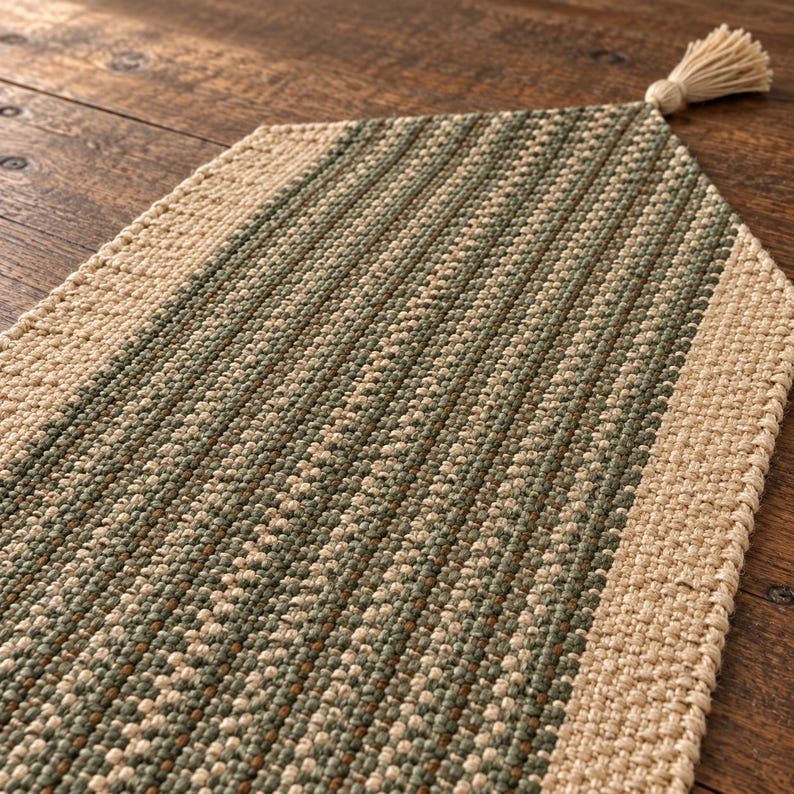 May include: A woven table runner featuring a central panel of sage green and beige textured fibers, bordered by a solid beige woven edge. The runner has a pointed end finished with a single beige tassel attached to the center point.