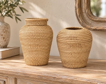 May include: Two light brown woven rattan vases with a tight, uniform grid pattern. The taller vase features a distinct neck and a braided rim, while the shorter vase has a rounded, bulbous shape with a smooth, open top opening.