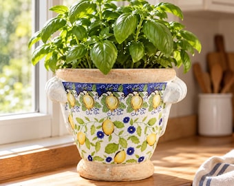 May include: A ceramic planter with a wide rim and two side handles, featuring a hand-painted pattern of yellow lemons, green leaves, and small blue flowers on a white background, containing a lush green basil plant with many leaves.
