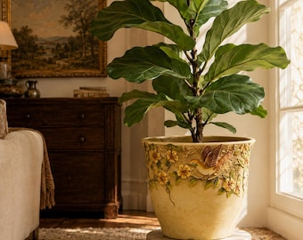 May include: A large fiddle leaf fig plant potted in a cream-colored ceramic planter. The planter features a raised, three-dimensional relief design of a small bird perched among yellow flowers and green vines encircling the upper exterior rim.