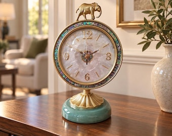 May include: A round table clock featuring a white stone face with gold numerals, surrounded by a border of iridescent abalone shell. A gold elephant figurine sits atop the frame, which is supported by a fluted gold pedestal and a smooth green base.