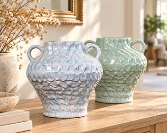 May include: Two ceramic vases with two handles each, featuring a textured, scale-like pattern on the exterior. One vase is finished in a glossy, iridescent light blue, while the other is a solid, soft sage green. Both have a wide, rounded body.