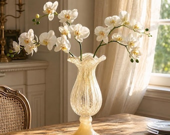 May include: A tall, clear glass vase with a textured, ribbed surface and a flared, ruffled rim, standing on a pedestal base. The vase contains several stems of white orchid flowers with yellow centers, arranged in a spreading, natural display.