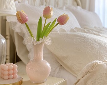 May include: A pale pink glass vase with a ruffled rim containing three artificial tulips with pink and yellow petals and green stems. Next to the vase sits a small, square, light pink bubble candle on a circular gold-rimmed white base.
