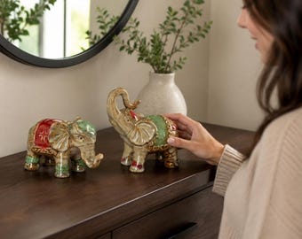 May include: Two ceramic elephant figurines with glossy finishes. One elephant features a red saddle and green accents, while the other has a green saddle and red accents. Both figures display intricate embossed patterns and are standing on a surface.