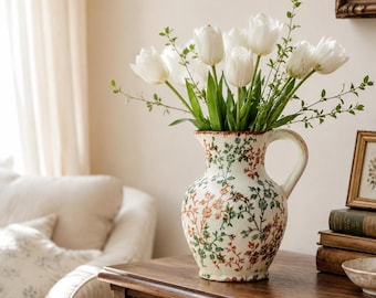 May include: A ceramic pitcher vase with a cream-colored base and a delicate botanical pattern in shades of green and orange. The pitcher features a curved handle and a wide rim, currently holding a large bouquet of fresh white tulips and greenery.