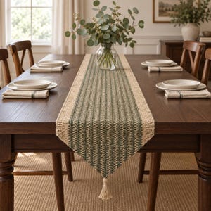Puede incluir: Un camino de mesa tejido rectangular con un patrón central de rayas verde oscuro y tostado con bordes en color tostado natural. El camino de mesa tiene un acabado texturizado y una borla decorativa que cuelga del extremo puntiagudo sobre una mesa.